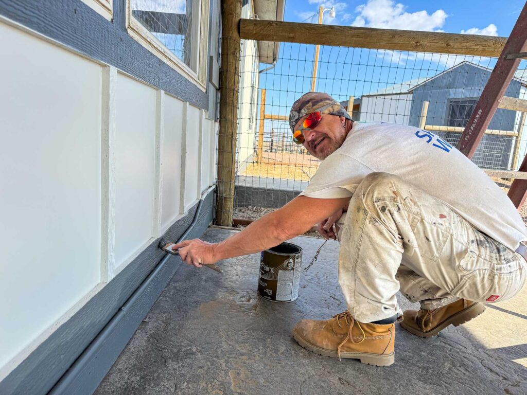 A painter working on the exterior trim of a house, with a can of paint placed behind him